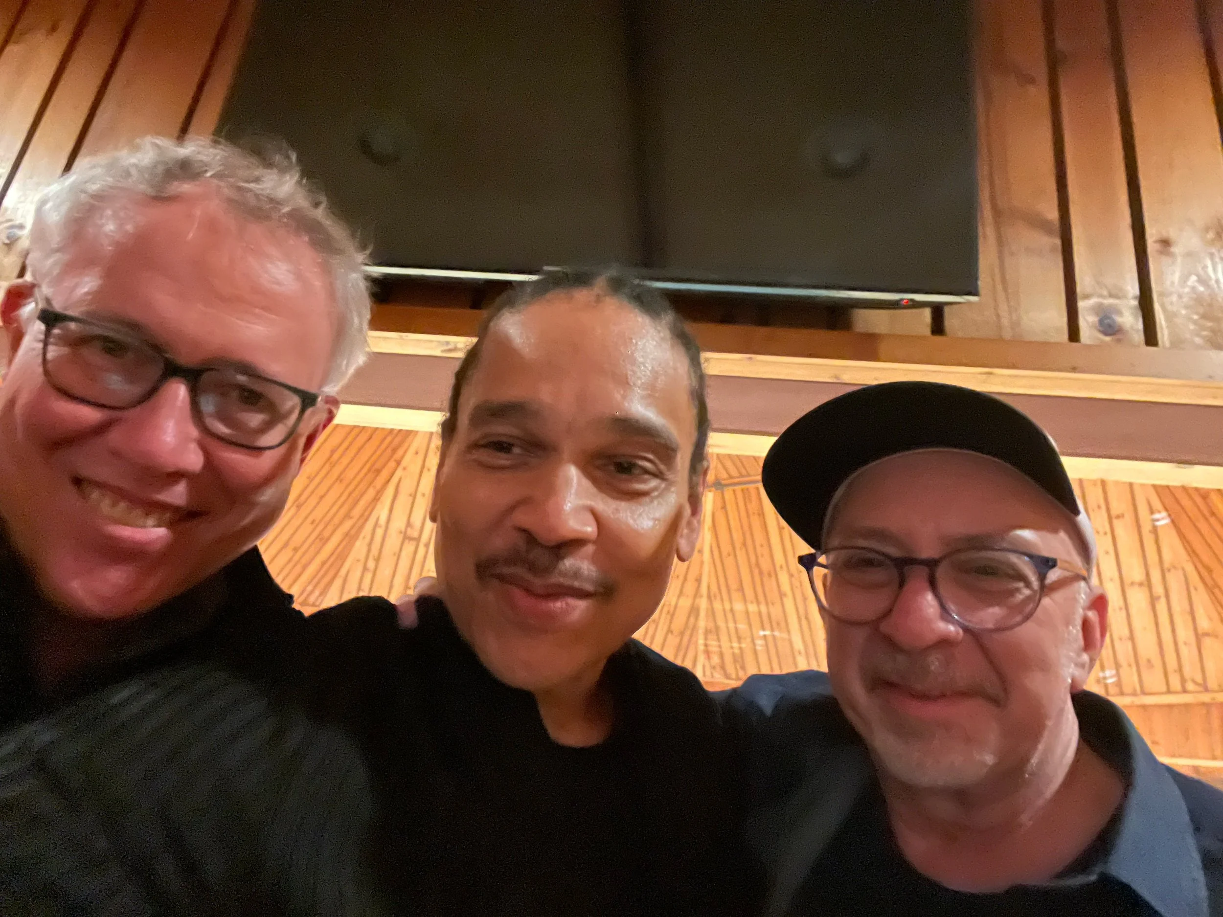 Three men taking a selfie together in a room with wooden walls and a large TV screen in the background. The man on the left has gray hair, glasses, and is smiling. The man in the middle has dark hair and is also smiling. The man on the right has gray hair, glasses, and is wearing a black cap.