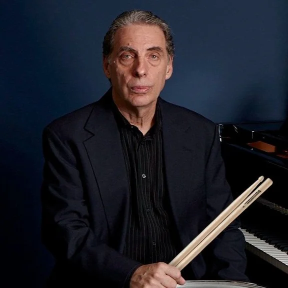 A man sitting at a piano holding drumsticks, wearing a dark blazer and black shirt, against a dark blue background.