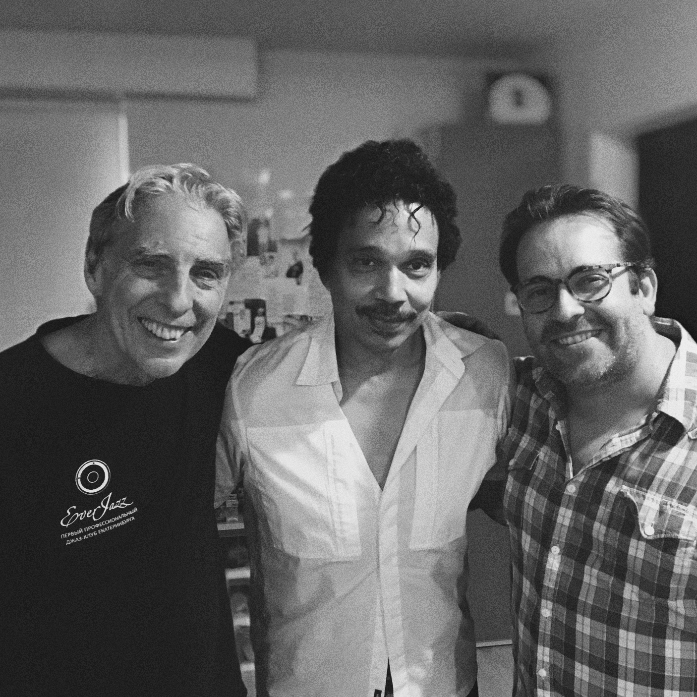 Three men smiling and posing together, one in a black T-shirt with a logo, another in a light-colored shirt, and the third in a plaid shirt, in a room with various objects in the background.