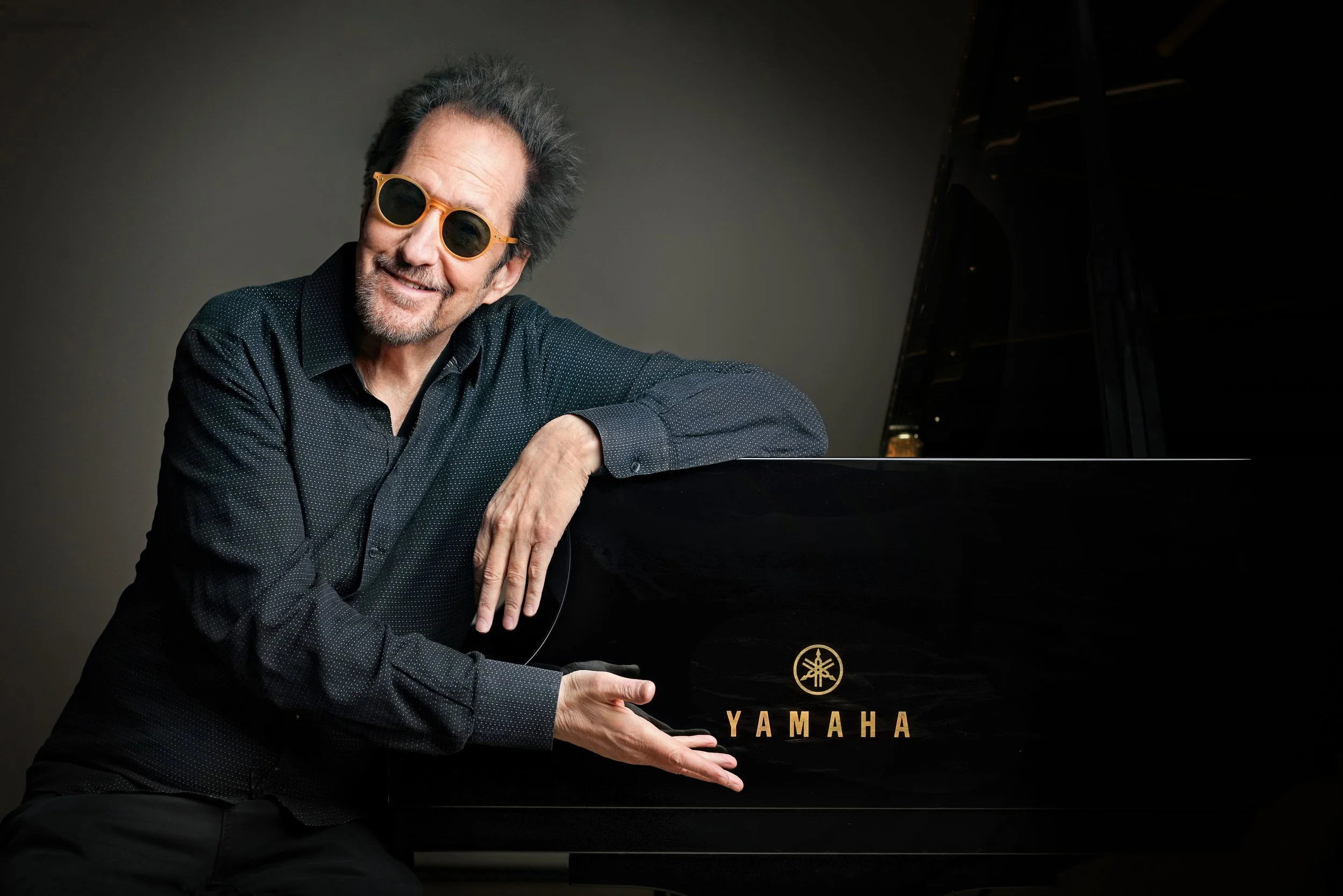 Man wearing sunglasses leaning on a black Yamaha grand piano.