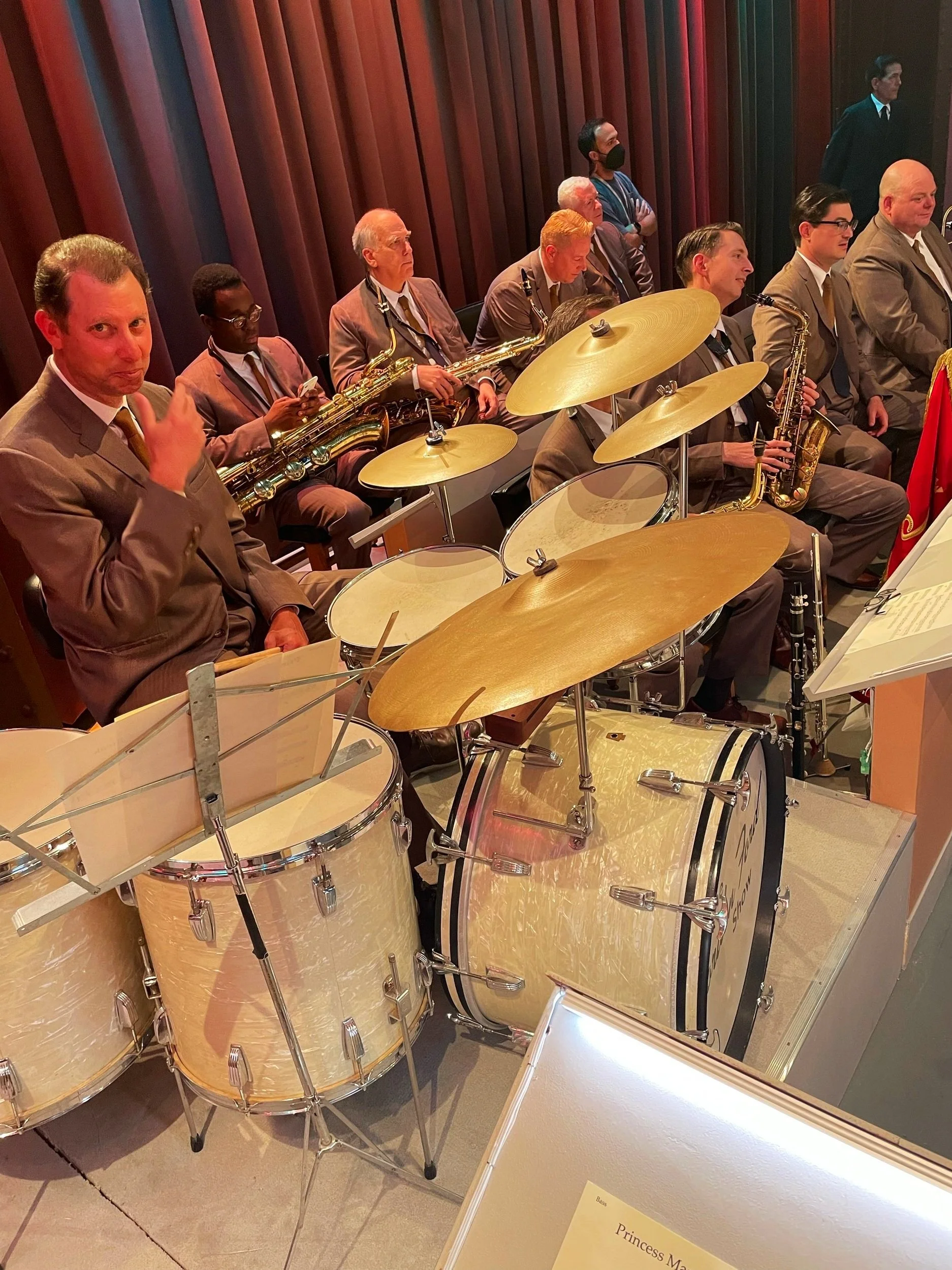 A group of musicians in suits seated with their instruments, including saxophones and drums, in an indoor setting with wooden walls. One person in the foreground is making a peace sign.
