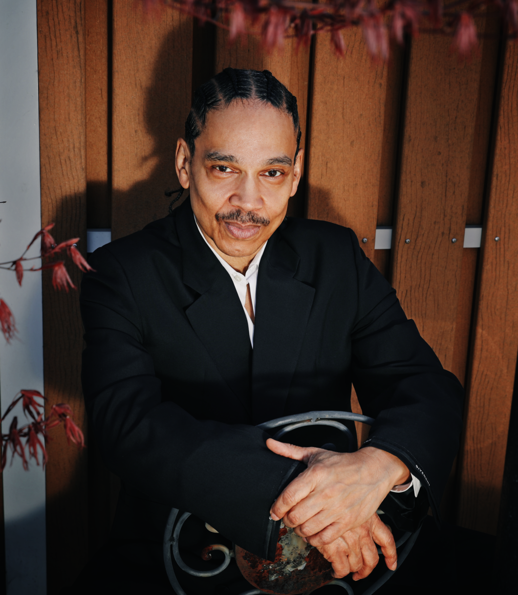 A man wearing a black suit and white shirt sitting against a wooden wall, holding a decorative spherical object, with red leaves in the foreground.