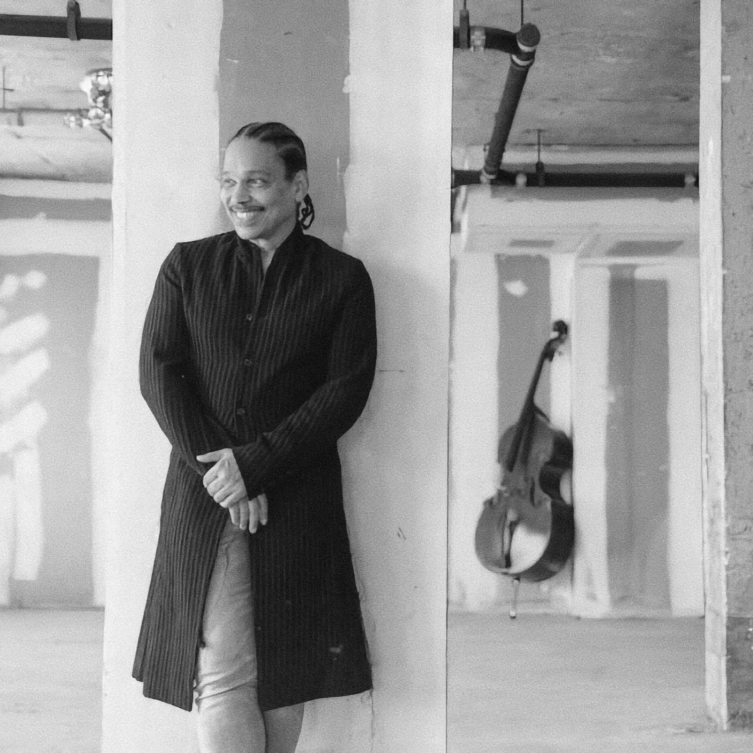 Mr. Dorsey, with slicked-back hair and earrings, wearing a long, striped black coat and light-colored pants, standing and smiling in a construction or studio space. There are exposed pipes and a guitar hanging on the wall behind her.