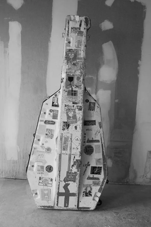 A human-sized, guitar-shaped object covered in various stickers, standing against an unfinished or textured wall.
