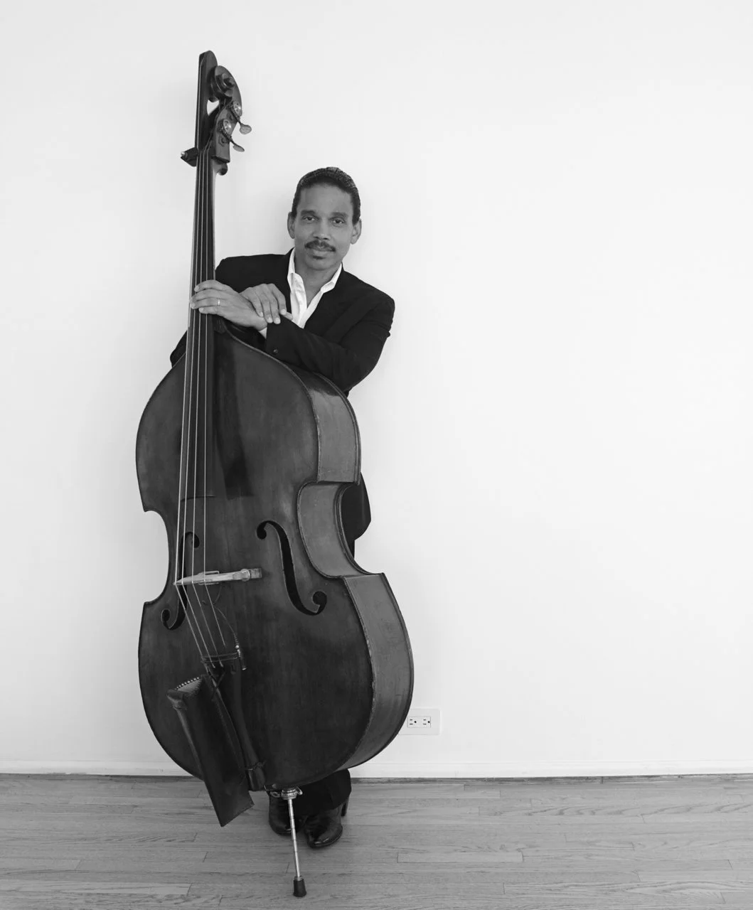 Man in a suit standing next to a large double bass in a room with a plain white wall.
