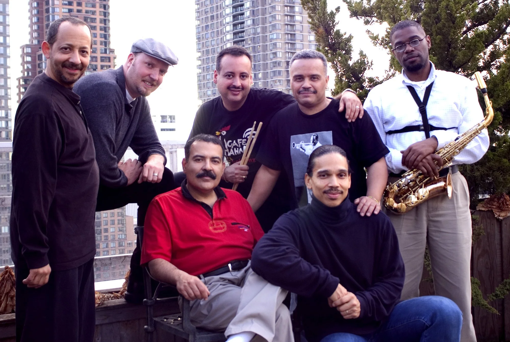 A group of seven men, some holding musical instruments, standing and sitting outdoors with city buildings and trees in the background.